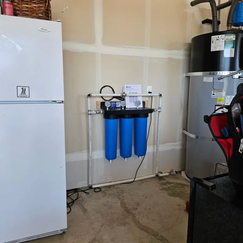 Three-stage blue whole-house water filtration system mounted in a garage beside an electric water heater
